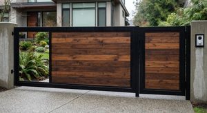 Modern automated driveway gate featuring dark metal framing and warm wooden horizontal slats, installed in front of a contemporary home with concrete pillars.